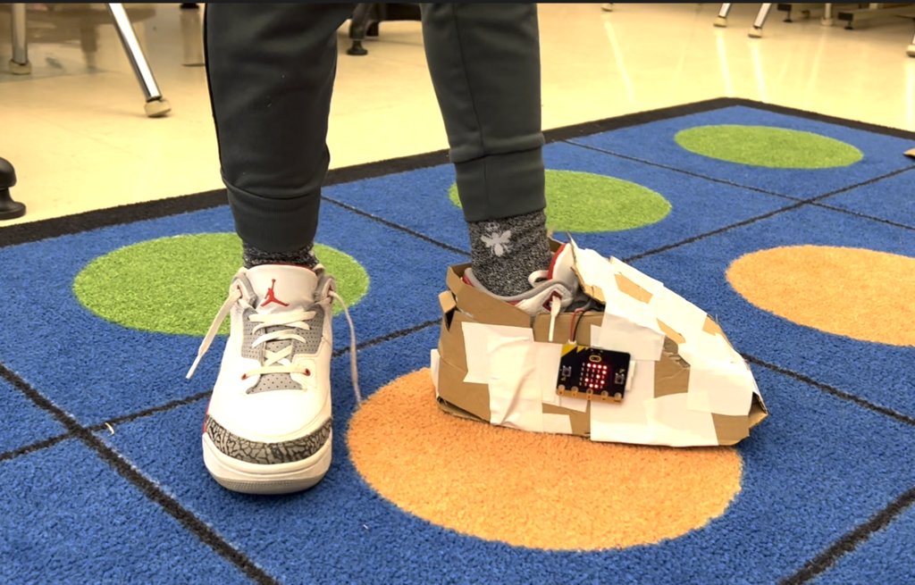 Student wearing a shoe made of cardboard and duct tape with a Micro:Bit step counter attached to the shoe.